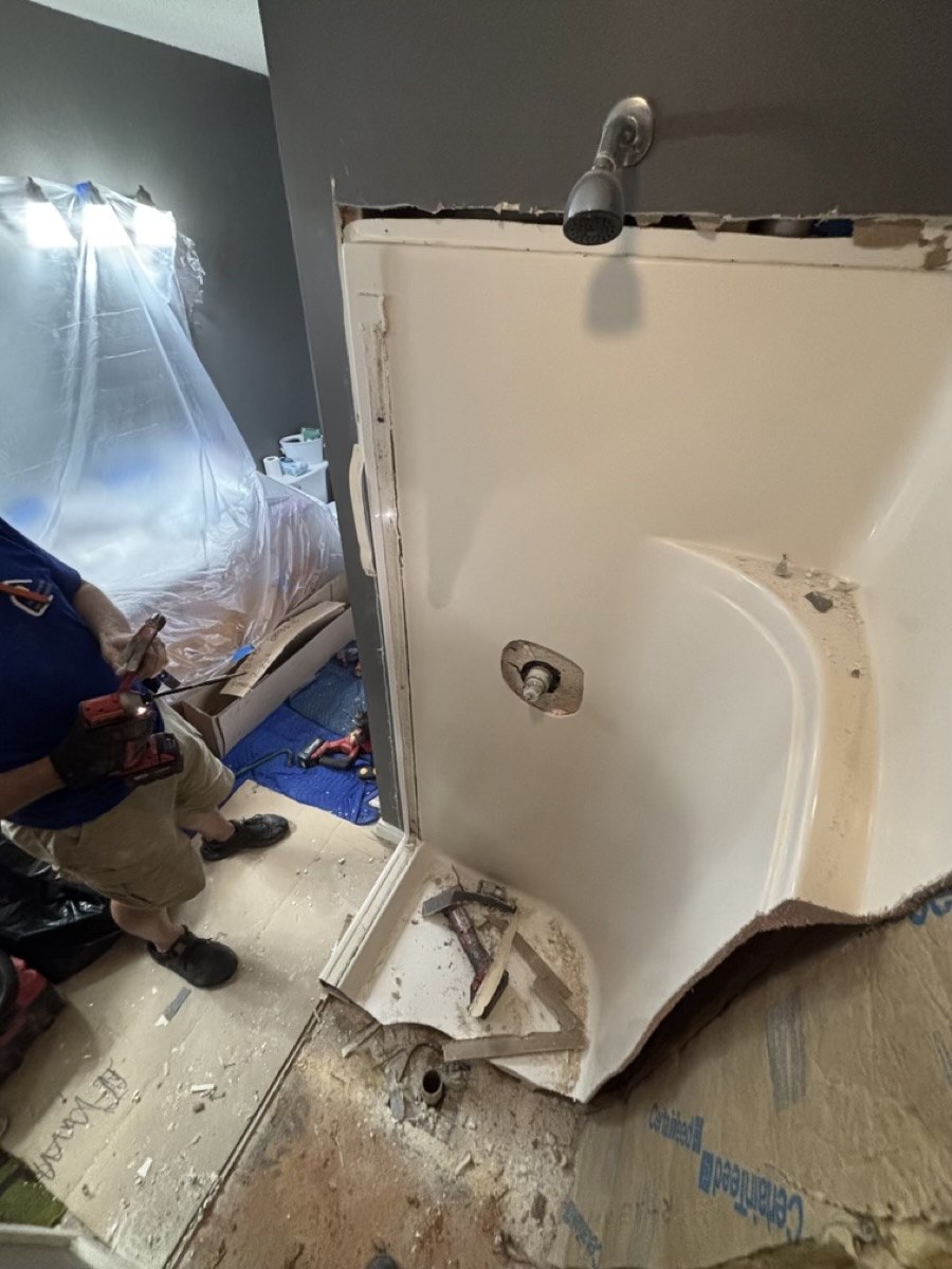 Before: a technician cutting out an old fiberglass bathtub surround during bathroom demolition, with plastic sheeting protecting the room.
