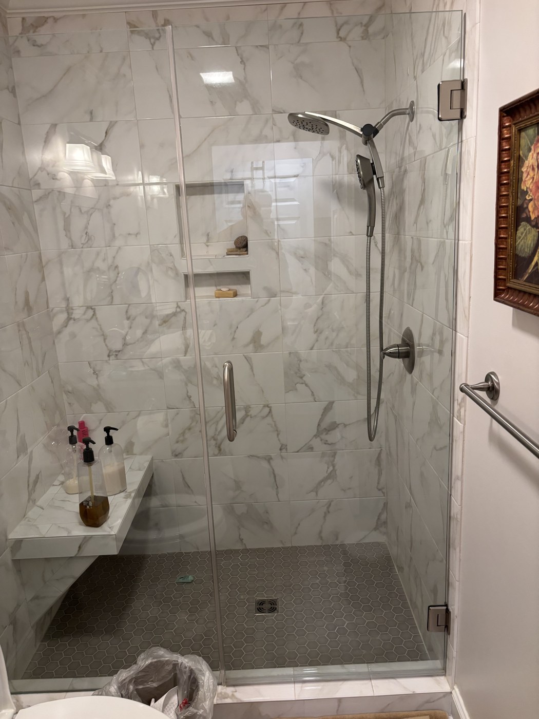 After: a finished marble-tiled walk-in shower with a glass door, built-in corner seat, recessed niche shelves, handheld shower wand, and a stainless support bar.